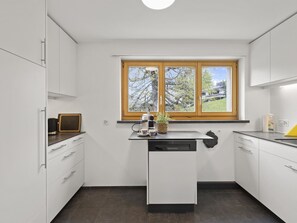 A modern kitchen with white cabinetry, a built-in dishwasher, a window with a view of trees, and a countertop with a coffee maker and a plant. - Bellavista 6 Appartement/Fewo, Bad, WC, 3 Schlafräume by Interhome (Parpan)