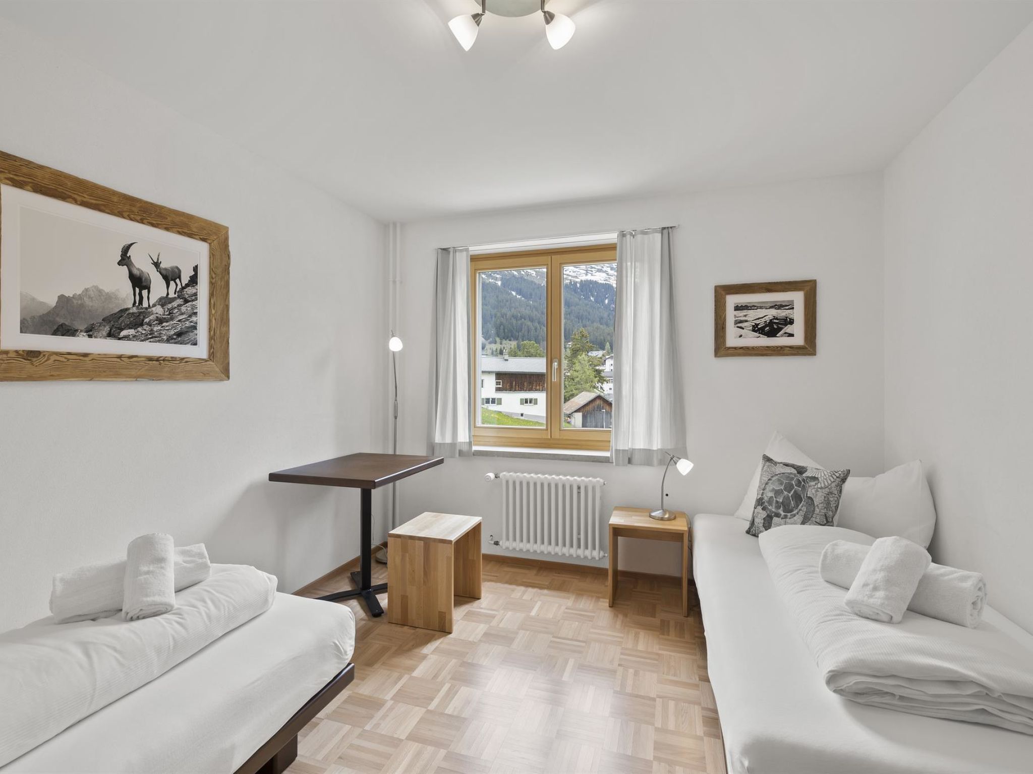 A room with two beds, a table, a radiator, and a window with a view of mountains.