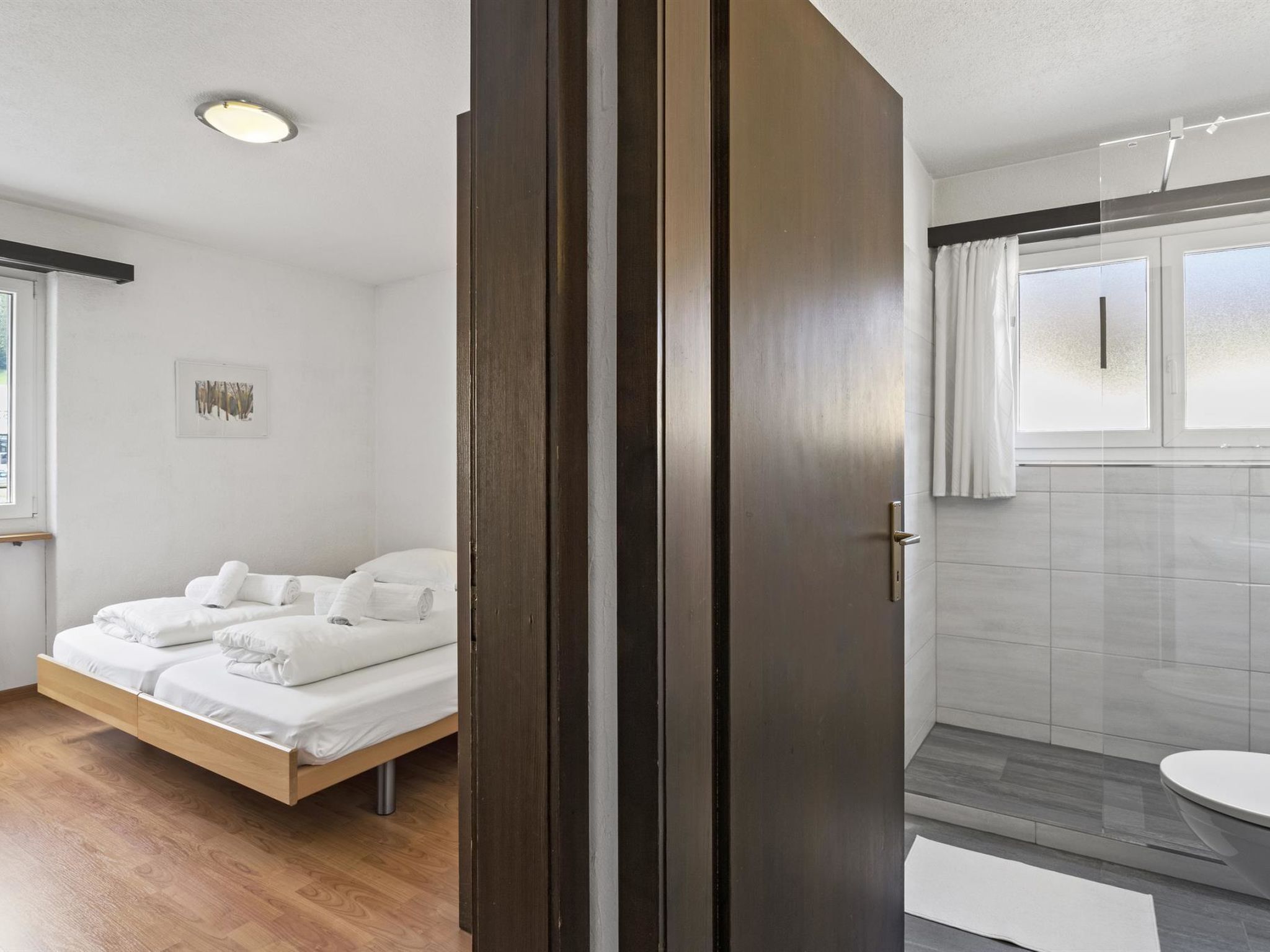Een moderne badkamer met een douche, toilet en een glazen deur die leidt naar een slaapkamer met een bed en een raam.