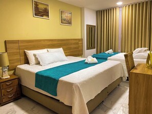 Habitación de hotel con dos camas, cabecera de madera, mesita de noche con lámpara y ventana con cortinas.