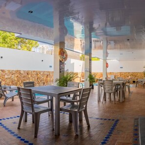 Un área de comedor al aire libre con mesas y sillas, un muro de piedra y una piscina al fondo.