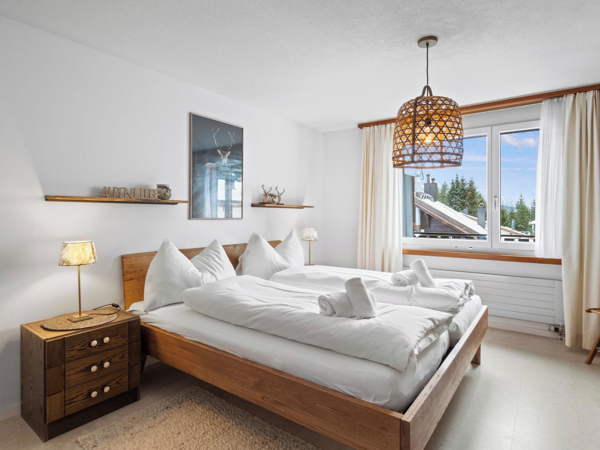 Een slaapkamer met een houten bed, een nachtkastje met een lamp, een wandplank met decoratieve objecten en een raam met gordijnen.