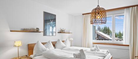 A bedroom with a wooden bed, a bedside table with a lamp, a wall-mounted shelf with decorative items, and a window with curtains.