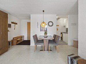 A modern dining area with a wooden table and grey upholstered chairs, a hanging light fixture, and a kitchen in the background. - A302 Appartement/Fewo A302 IH by Interhome (Lenzerheide)