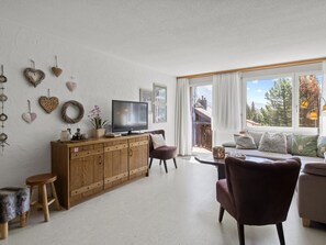 A living room with a wooden cabinet, a television, a sofa, and a view of a mountain landscape through large windows. - A302 Appartement/Fewo A302 IH by Interhome (Lenzerheide)