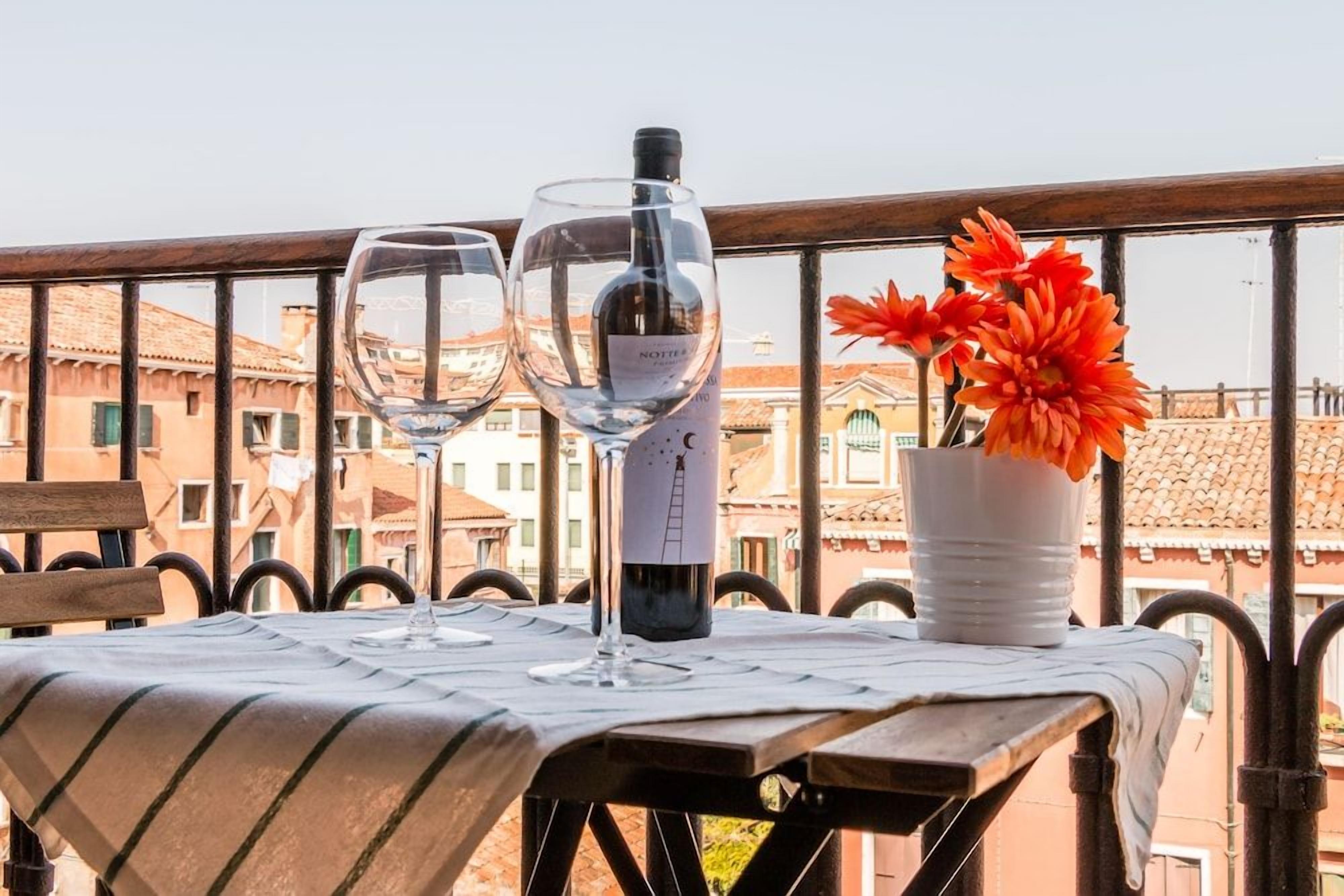 A table with two wine glasses and a bottle, a white vase with orange flowers, and a view of buildings in the background.