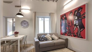 A modern living room with a sofa, a dining table with chairs, a clock, and a large abstract painting.