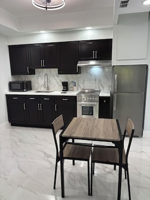 A modern kitchen with dark cabinets, a stainless steel refrigerator, a built-in oven, and a wooden dining table with chairs. - Avalon Resort (Deerfield Beach)