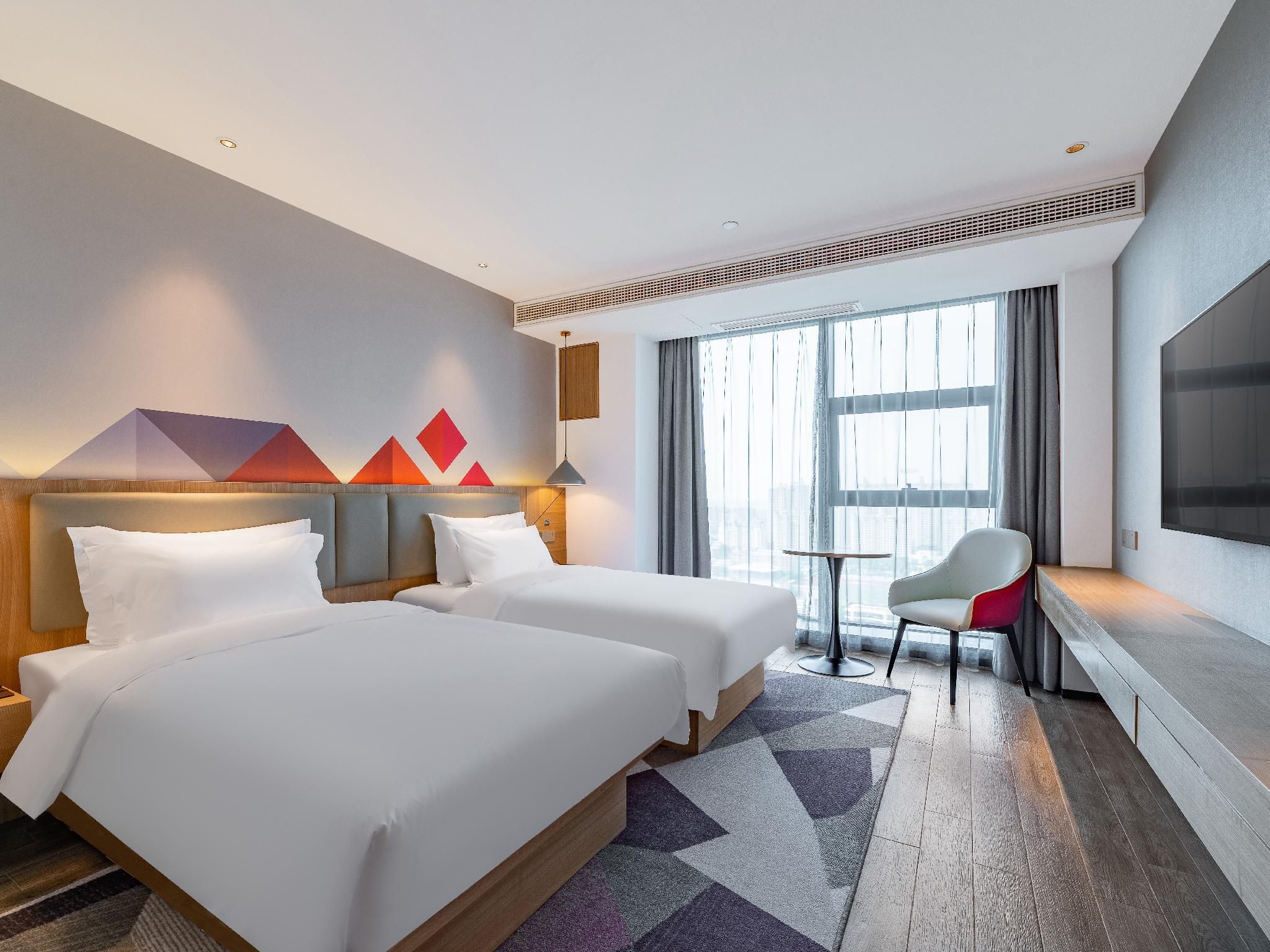 A hotel room with two beds, a TV, a desk, and a large window.