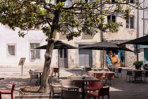 An outdoor cafe with tables and chairs, a tree, and a mural on the building. - Hotel & Restaurant Des Basses Pyrenees (Bayonne)