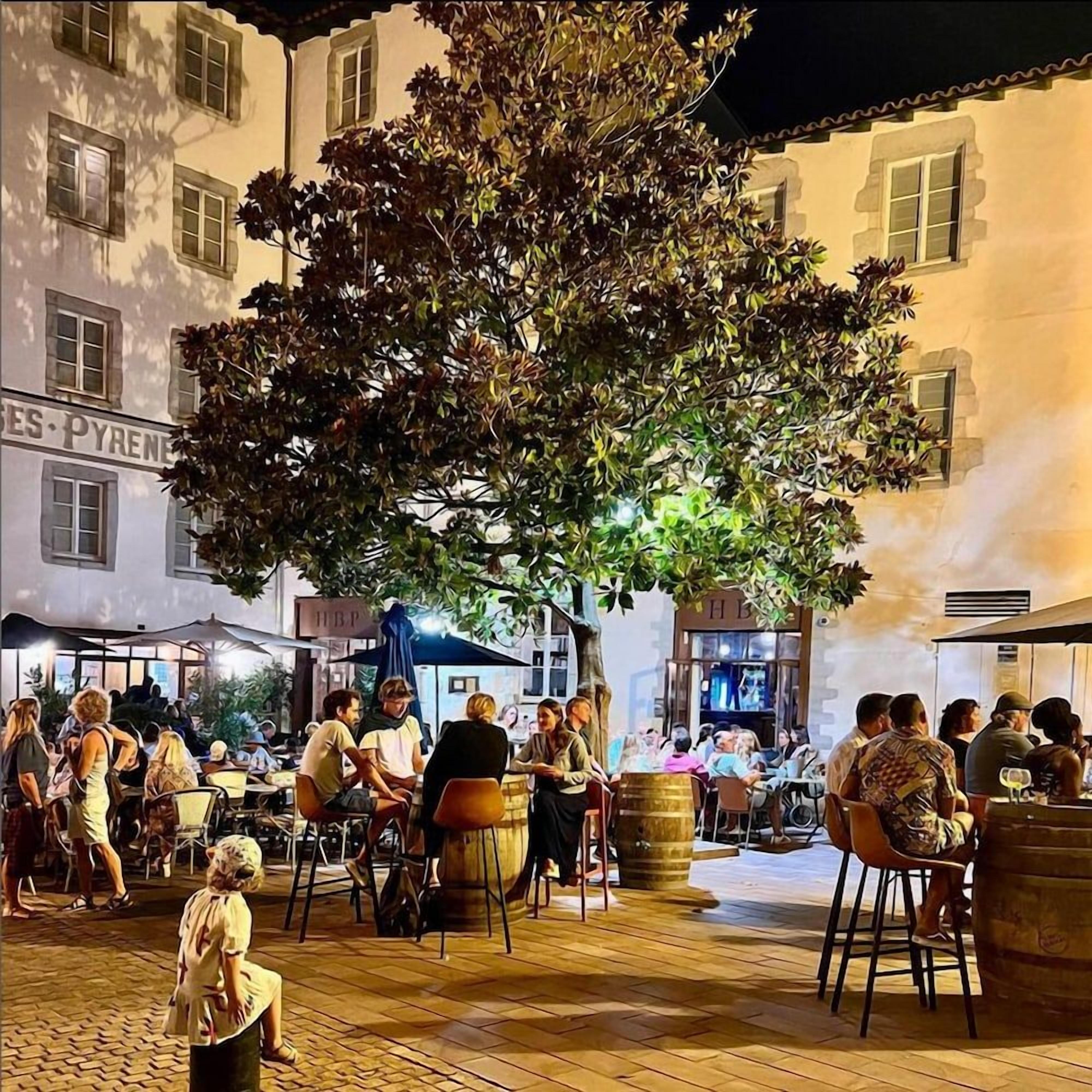Un espace de café en plein air, avec des tables et des chaises, entouré de bâtiments portant des enseignes.