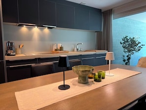 A modern kitchen with dark cabinets, a coffee machine, and a dining area with a table, chairs, and a lamp. - Eden Alpine by Arosa Holiday Appartement/Fewo, Badewanne, 2 Schlafräume by Interhome (Arosa)