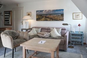 A cozy living room with a sofa, armchair, and a table. There is a painting on the wall and a lamp on a side table. - Comfortable apartment in Keitum with a beach chair on Kampen Beach (Sylt)