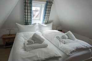A neatly made twin bed with white linens and rolled towels, a bedside table with a lamp, and a window with blue and white checkered curtains. - Comfortable apartment in Keitum with a beach chair on Kampen Beach (Sylt)