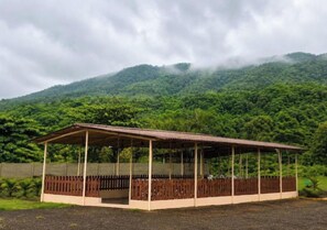 A covered pavilion with a wooden fence, set against a backdrop of a lush green hill.