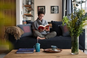 Eine Person sitzt auf einem dunklen Sofa, mit einem Buch, einer Kaffeekanne und einer Tasse auf einem Holztisch.