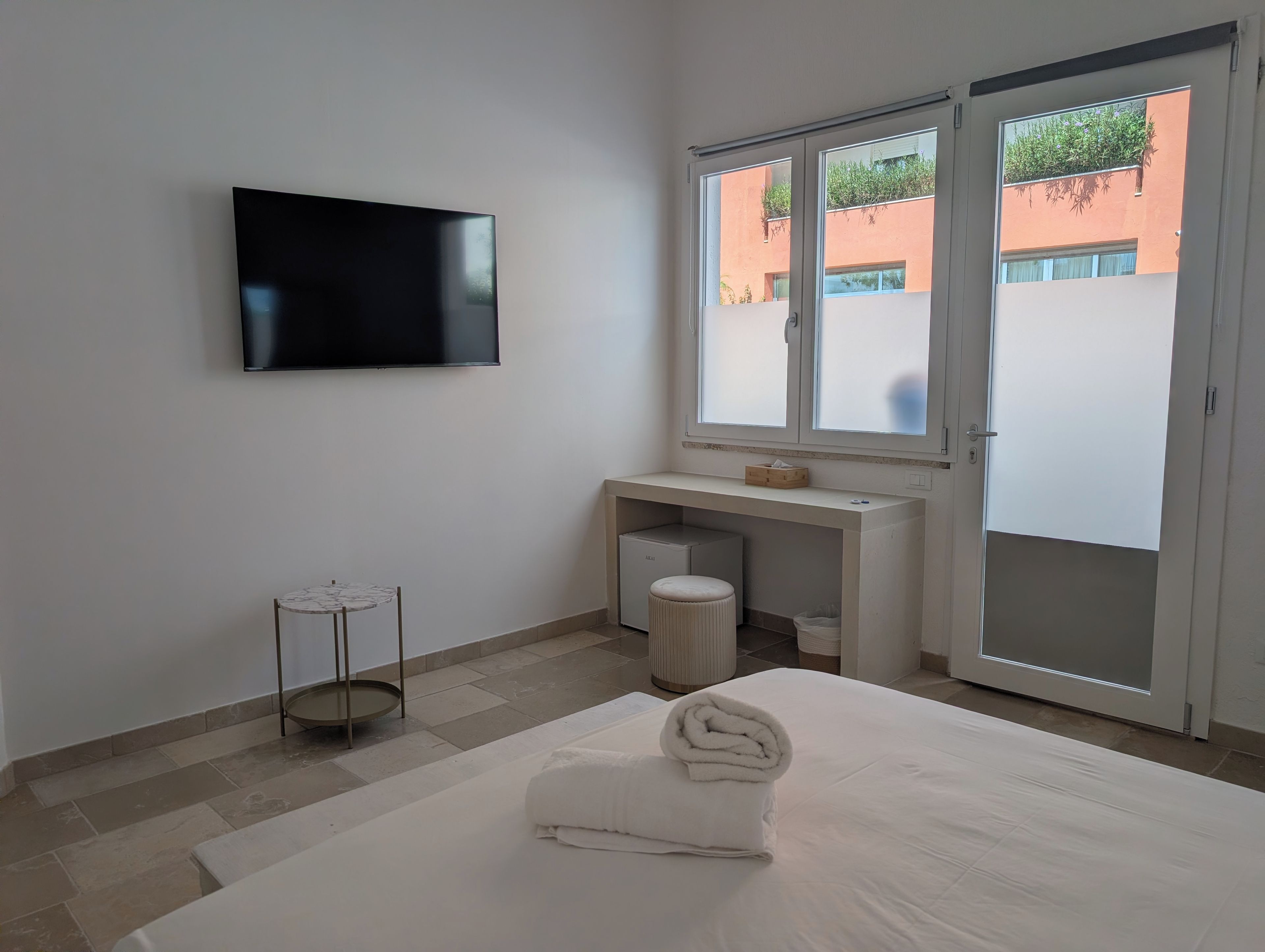 Ein Hotelzimmer mit einem Bett, einem an der Wand montierten Fernseher, einem kleinen runden Tisch, einem Fenster mit Blick auf Gebäude und einer Schiebetür.