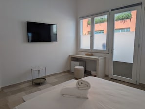 A hotel room with a bed, a TV mounted on the wall, a small round table, a window with a view of buildings, and a sliding door. - B&B Boutique Miramare (Lido Marini)