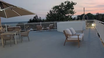 A rooftop terrace with outdoor seating, a view of the sea, and a sunset in the background.