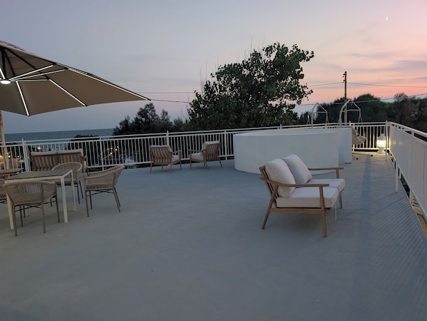 A rooftop terrace with outdoor seating, a view of the sea, and a sunset in the background.