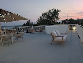 A rooftop terrace with outdoor seating, a view of the sea, and a sunset in the background.
