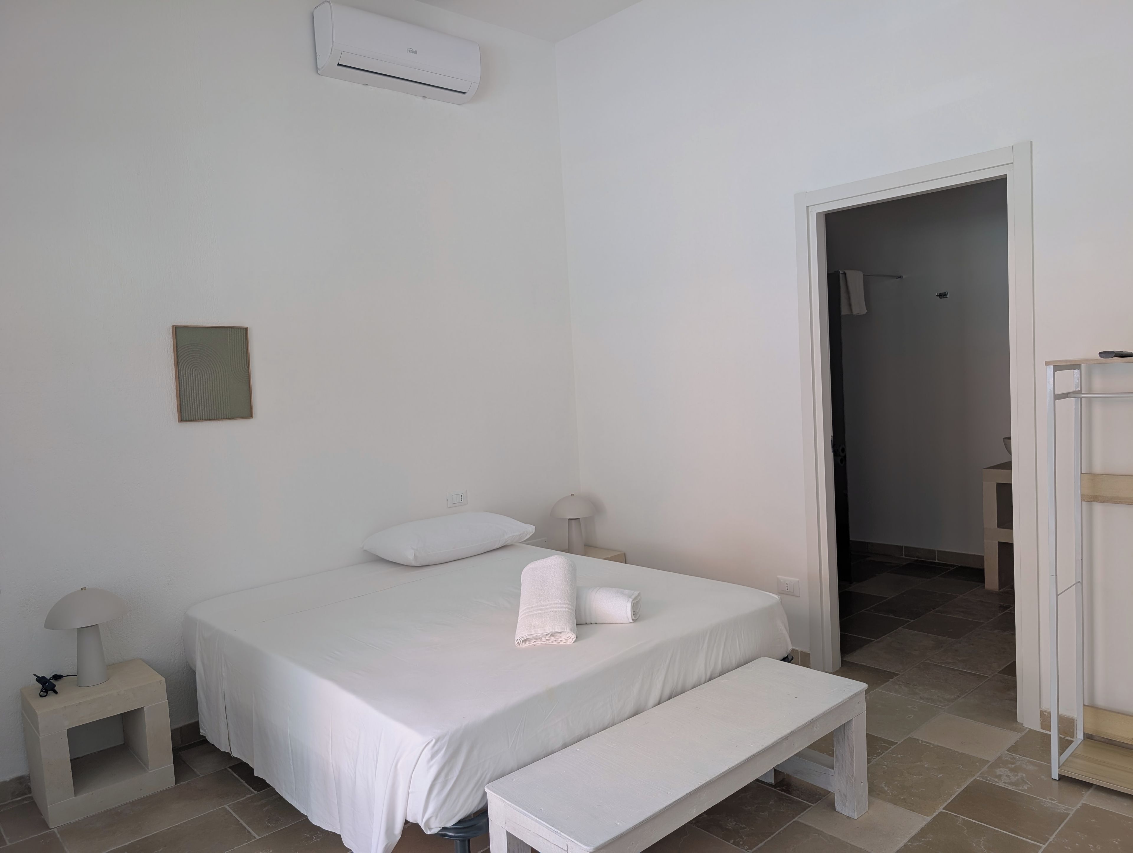 Un dormitorio con cama, mesita de noche, banco y unidad de aire acondicionado.