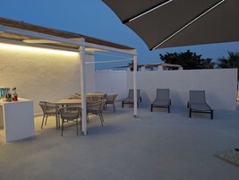 An outdoor patio with a white wall, a table with chairs, and a bar area with bottles.