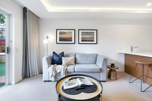 A modern living room with a grey sofa, a coffee table with a magazine, two framed pictures on the wall, and a kitchen area with a bar stool. - Cairns Central, your modern Oasis with pool (Cairns City)