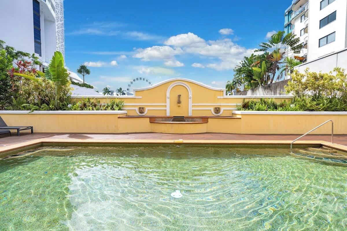Una piscina cristalina con un muro amarillo, un edificio blanco con balcones y plantas tropicales.