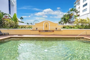 Une piscine aux eaux cristallines avec un mur jaune, un bâtiment blanc doté de balcons et des plantes tropicales.