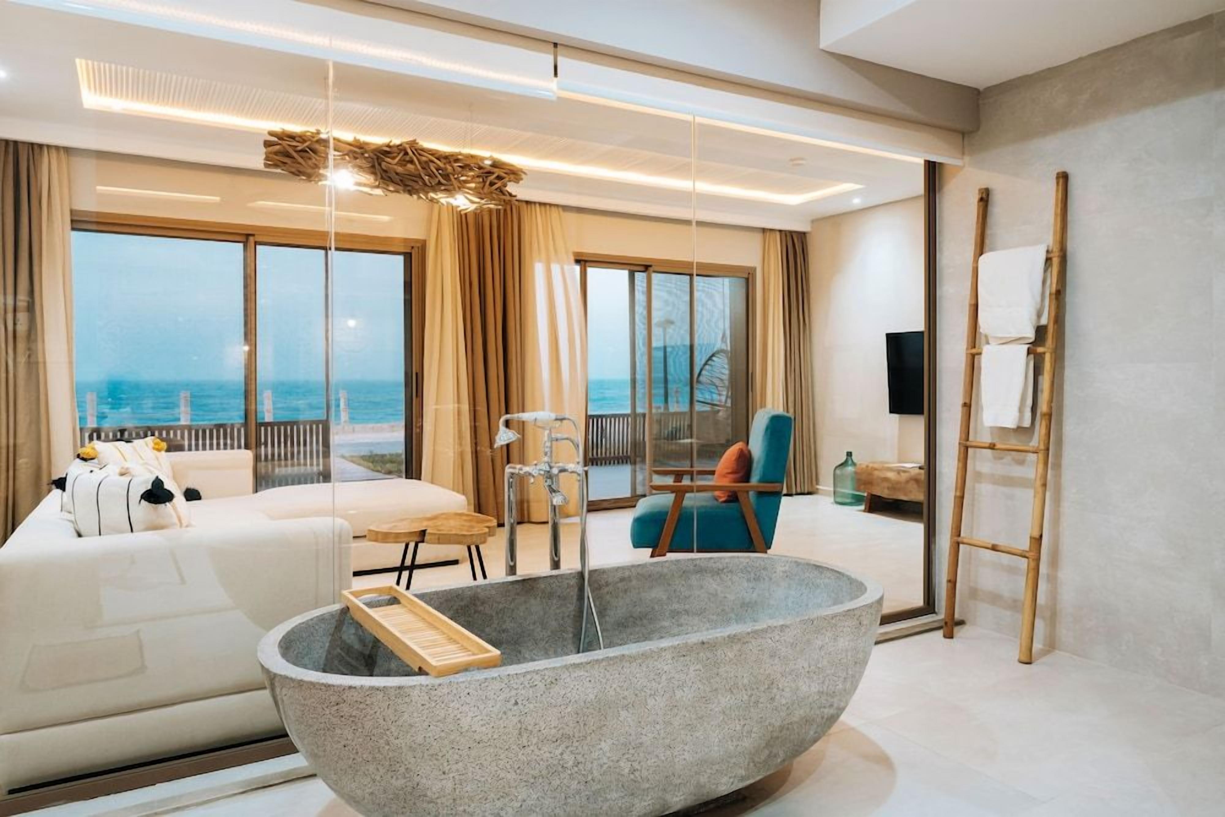 A modern hotel room with a large stone bathtub, a sofa, a dining area, and a view of the sea.