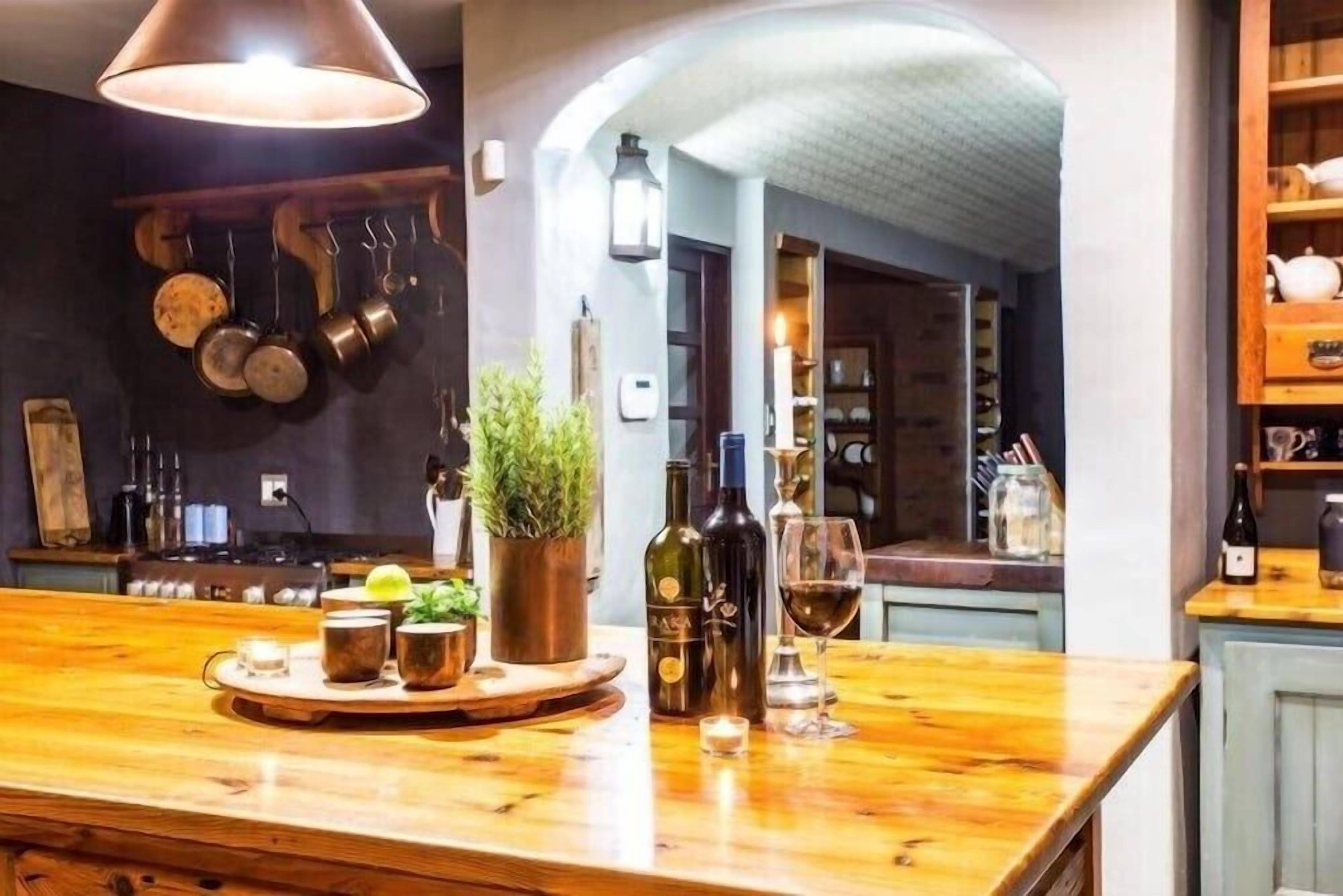 A bar area with a wooden counter, bottles, glasses, and a hanging pot rack.