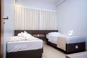 A hotel room with two beds, each with a duvet and pillows, and a window with sheer curtains.