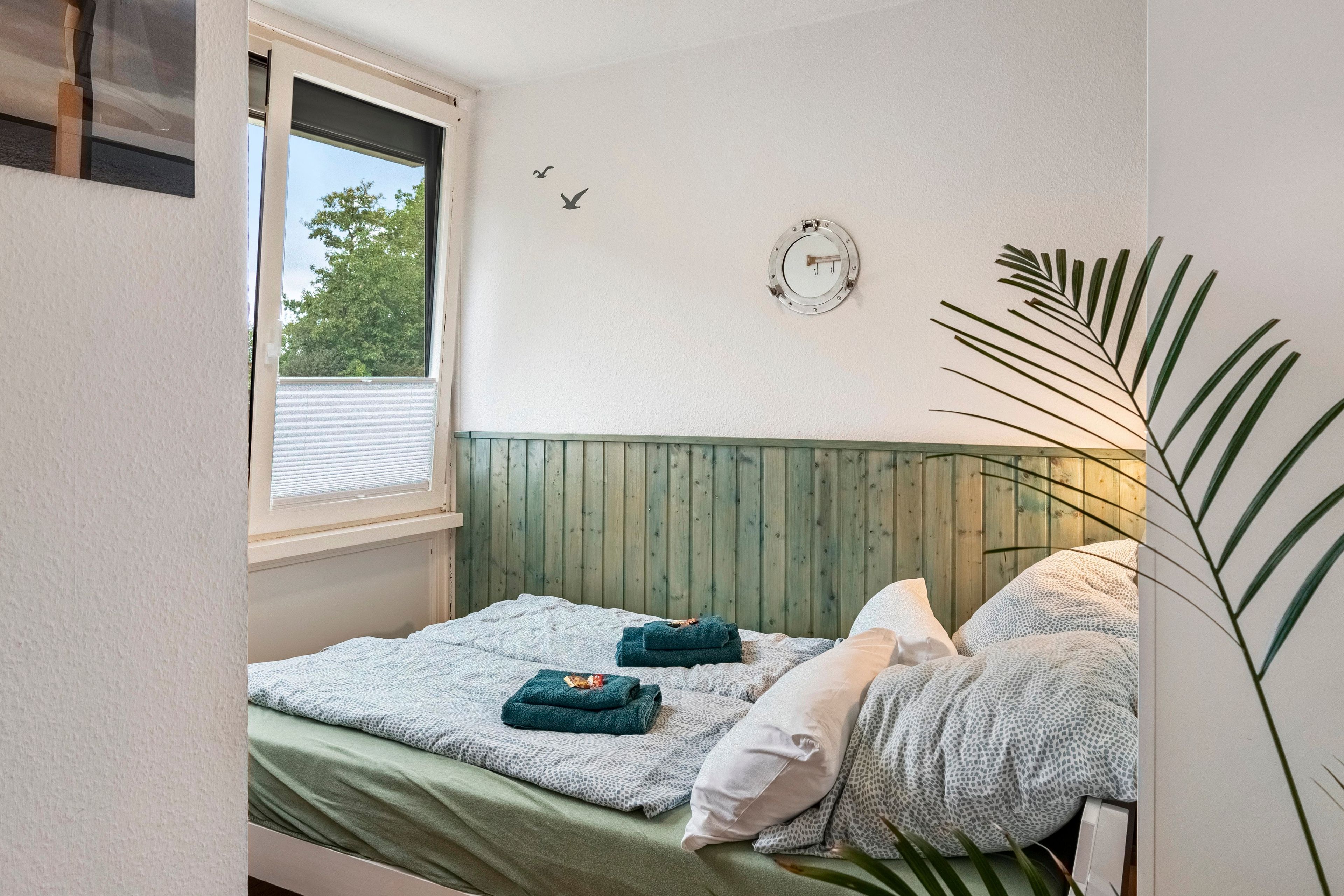 A bedroom with a bed, a window, a wooden headboard, a wall clock, and a plant.