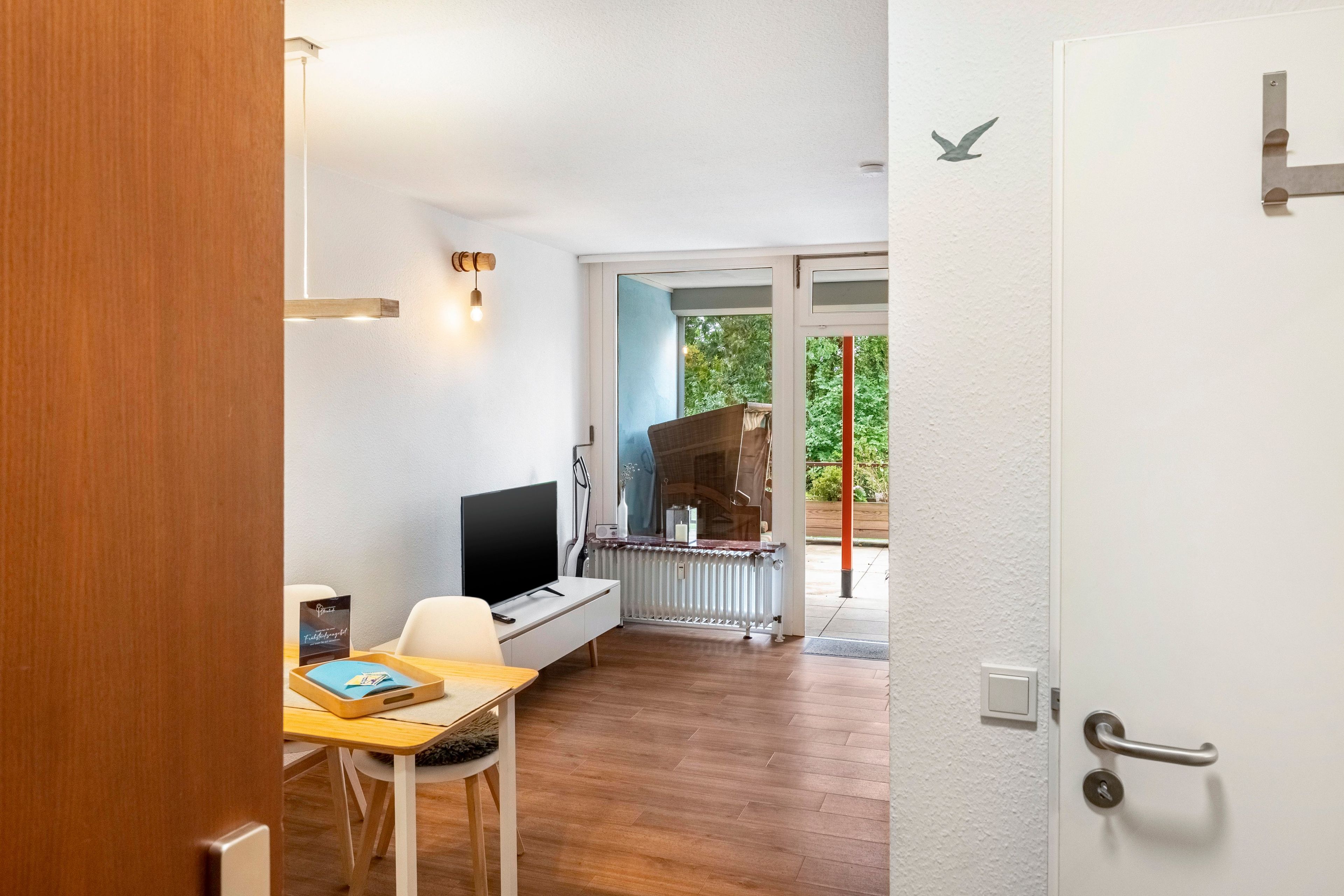 Ein Zimmer mit Holzboden, einer weißen Tür, einem Fernseher auf einem weißen Tisch und einem Fenster mit Blick auf grüne Vegetation.