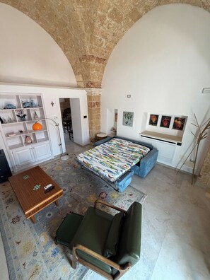A room with a bed, a sofa, a small table, and a chair. There is a bookshelf and a lamp. - Stella del Salento – Historic residence with a private terrace (Tricase)