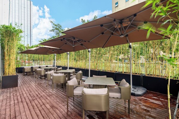 An outdoor seating area with tables and chairs, covered by umbrellas, and surrounded by bamboo plants. - Home2 Suites by Hilton Shenzhen Nanshan Science & Technology Park (Shenzhen)