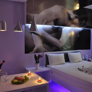 A modern hotel room with a large flat-screen TV, a bed with white bedding, a bedside table with a vase of flowers, and a small table with a candle and a glass of water. - Miragem Motel (Rio Verde)
