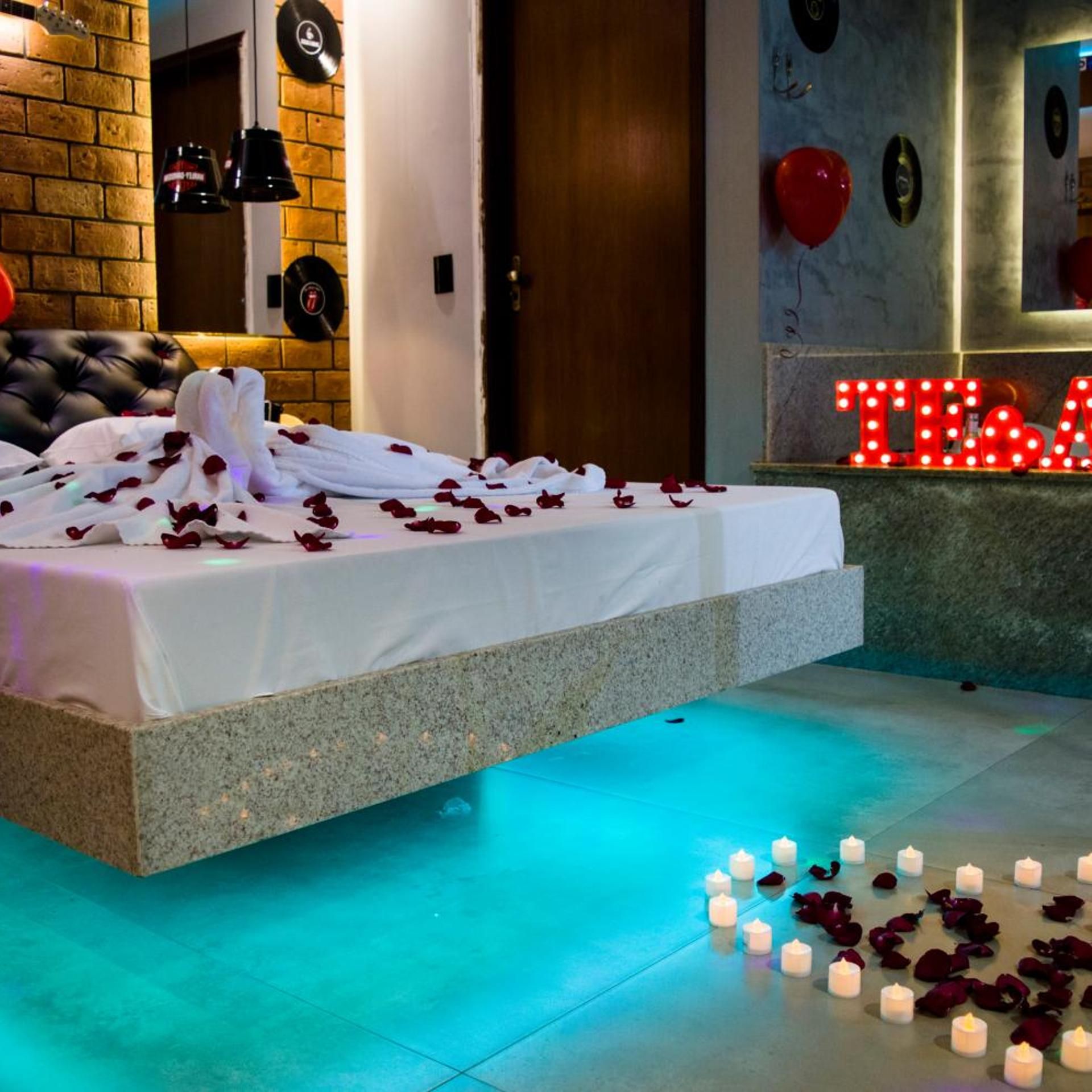 Quarto moderno com cama, pétalas de rosa, velas e um letreiro neon.