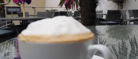 Una taza de café sobre una mesa, con vista a una terraza al aire libre y sombrillas.