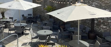 An outdoor patio area with round tables and chairs, two large umbrellas, and a brick wall in the background.