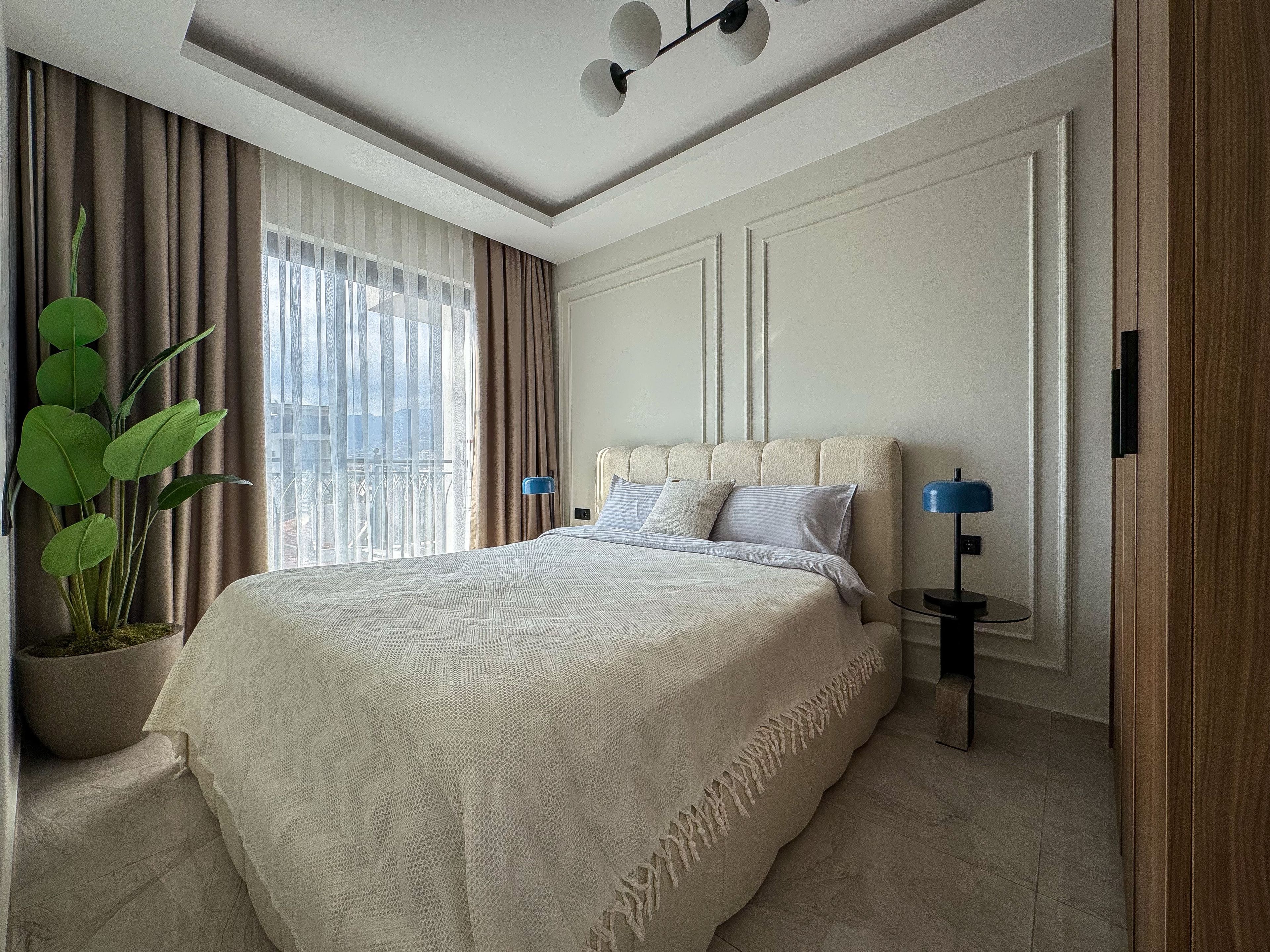 A modern bedroom with a large bed, a potted plant, and a view through the window.