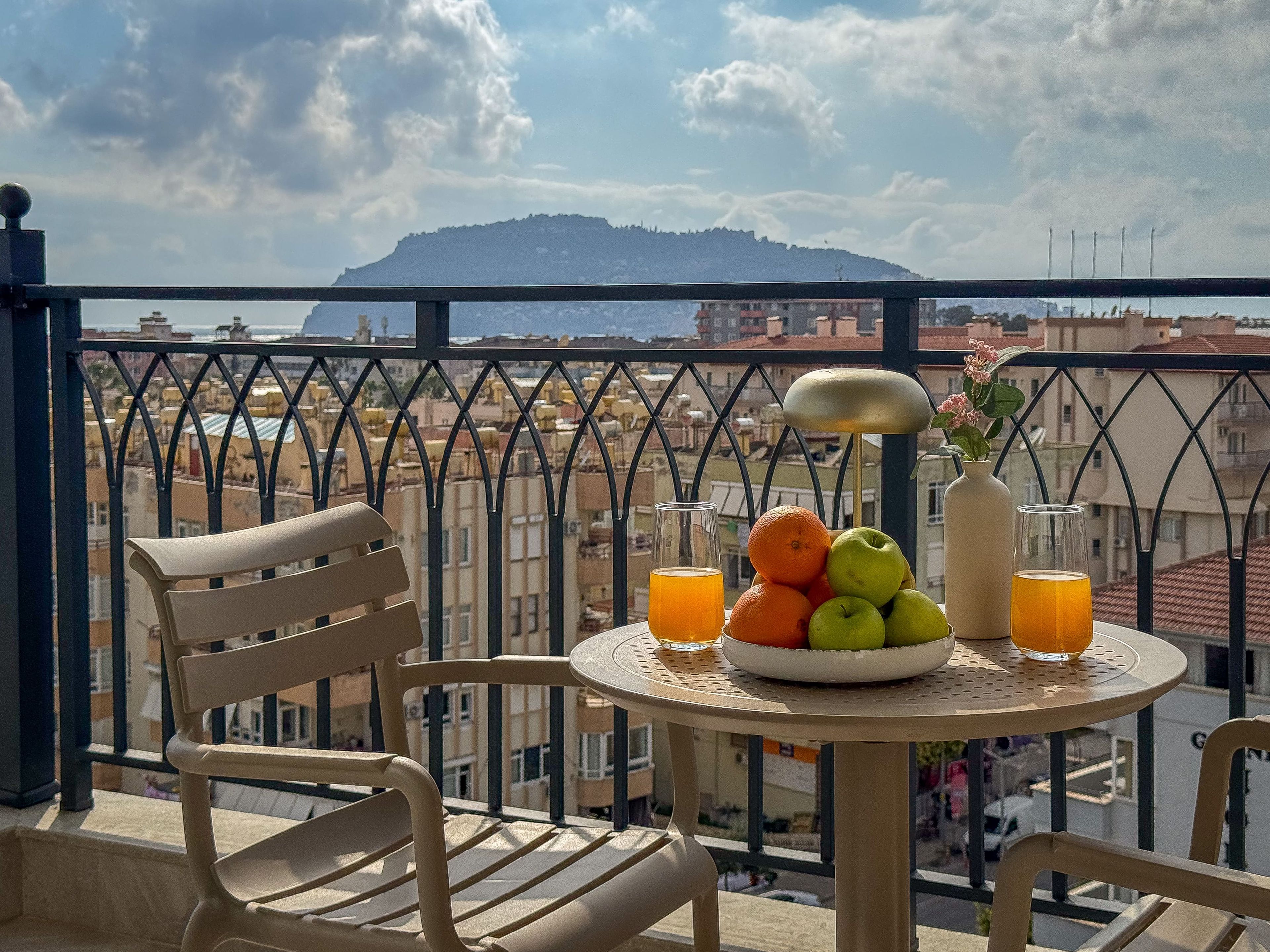 Un balcon avec une table préparée pour le petit-déjeuner, une chaise, un vase rempli de fleurs et une vue sur le paysage urbain et les montagnes.