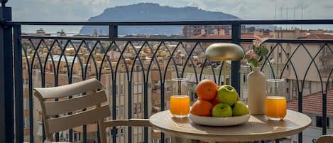 Un balcon avec une table préparée pour le petit-déjeuner, une chaise, un vase rempli de fleurs et une vue sur le paysage urbain et les montagnes.
