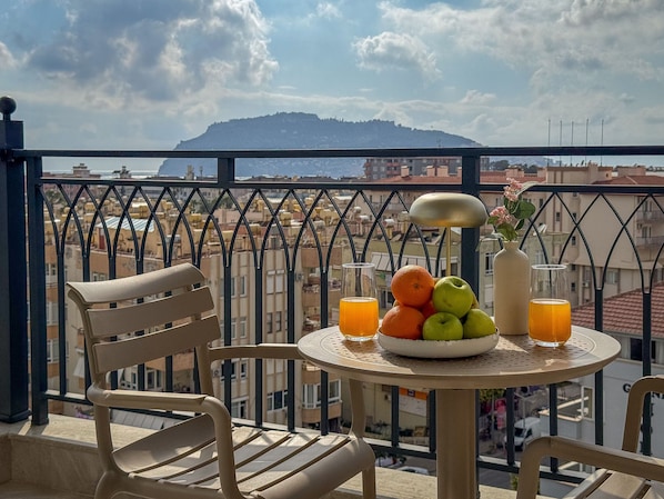 A balcony with a table set for breakfast, a chair, a vase with flowers, and a view of the cityscape and mountains.