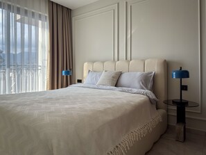 A hotel room with a large bed, two bedside lamps, a view through sheer curtains, and a cityscape.