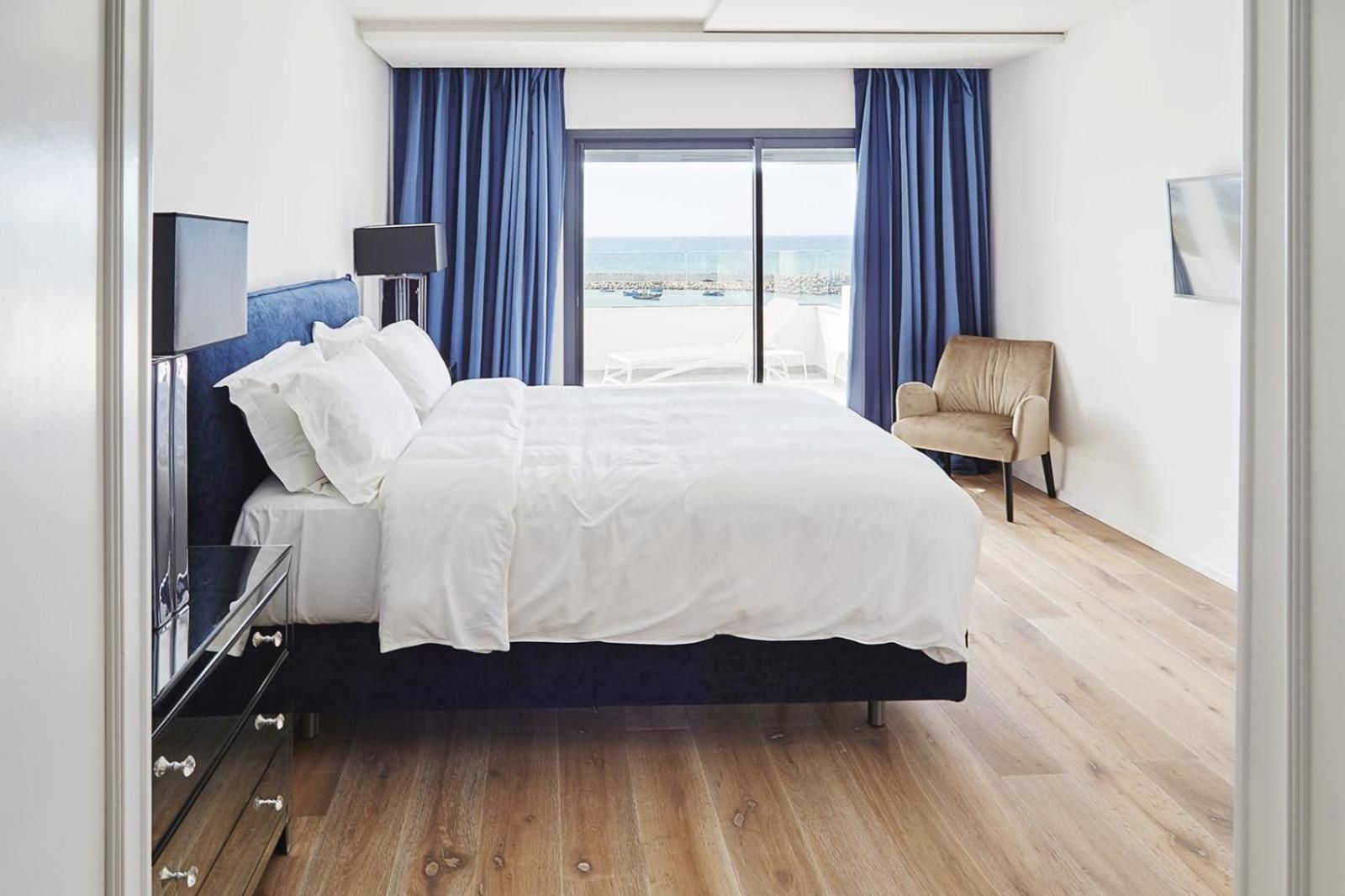 A hotel room with a bed, a TV, a chair, and a view of the sea.