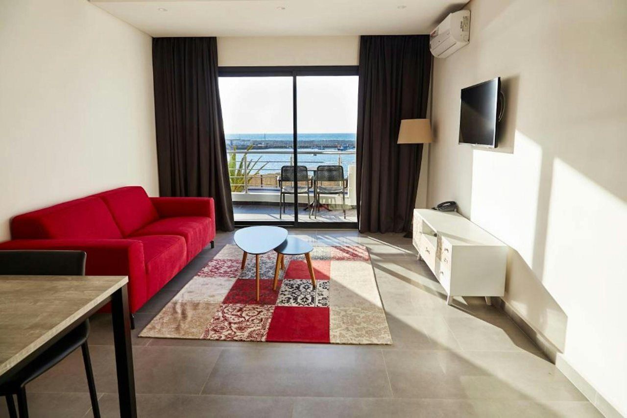 A living room with a red sofa, a small table, a TV mounted on the wall, and a view of the sea through large windows.
