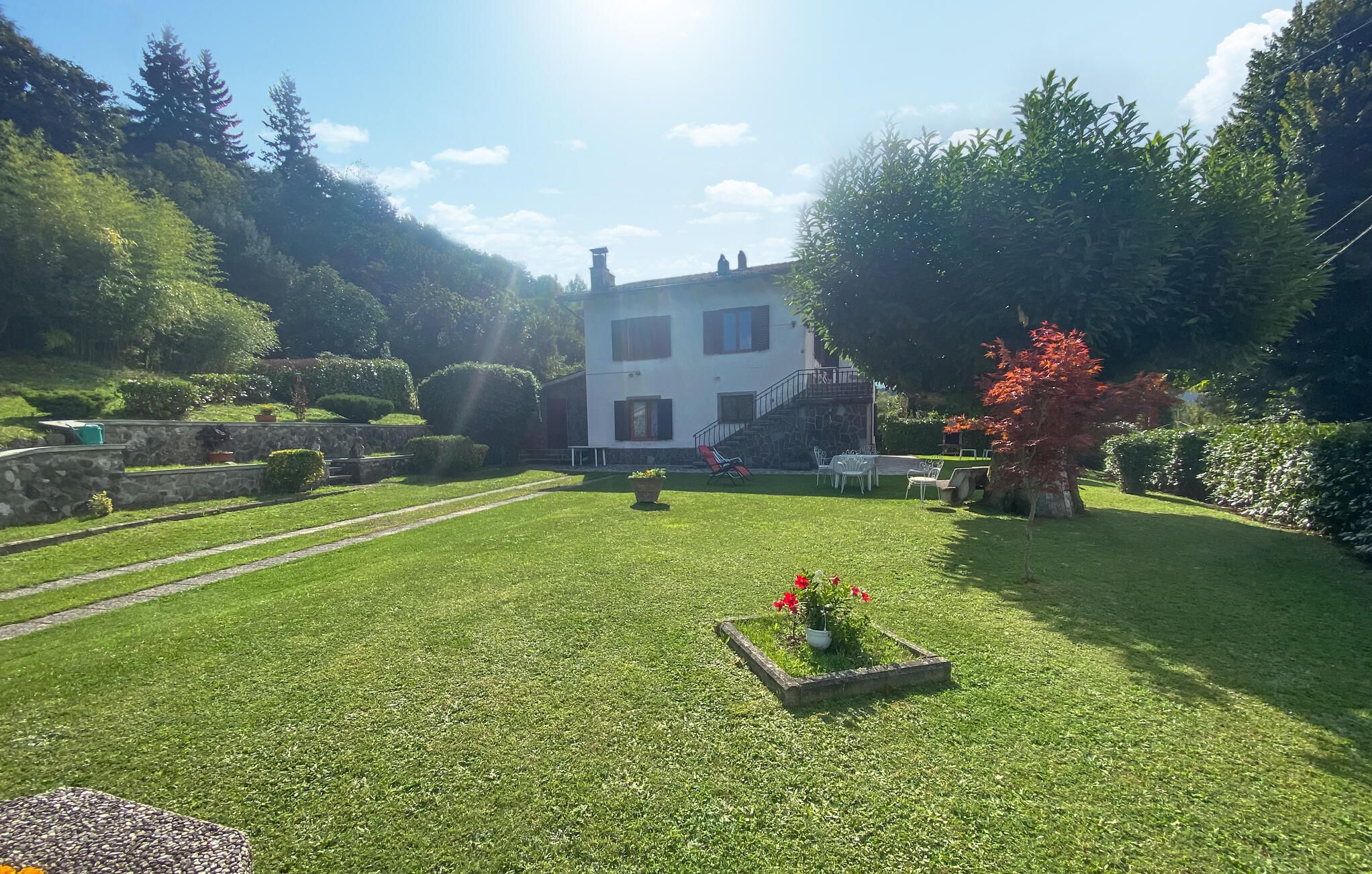 Uma casa de dois andares com uma escadaria de pedra, gramado e jardim com uma árvore de folhas vermelhas.