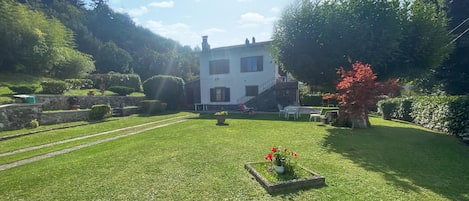 Uma casa de dois andares com uma escadaria de pedra, gramado e jardim com uma árvore de folhas vermelhas.
